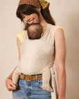 A woman wearing a cream and beige striped baby carrier wrap with a matching headscarf, carrying a baby against her chest. She is wearing a yellow top, blue jeans, and a brown belt, set against a plain beige studio background.