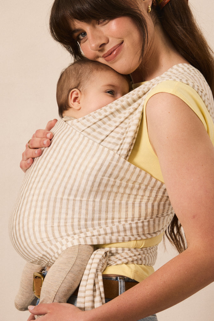 A woman wearing a beige and white gingham baby wrap carrier with a baby inside, styled over a yellow top against a plain beige background.