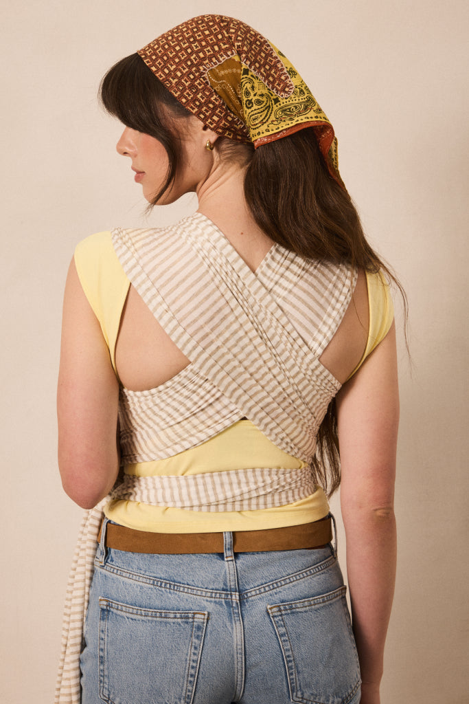 A woman shown from the back wearing a yellow sleeveless top with a beige and white striped wrap-style back panel, blue jeans, and a patterned brown headscarf, against a plain beige studio background.