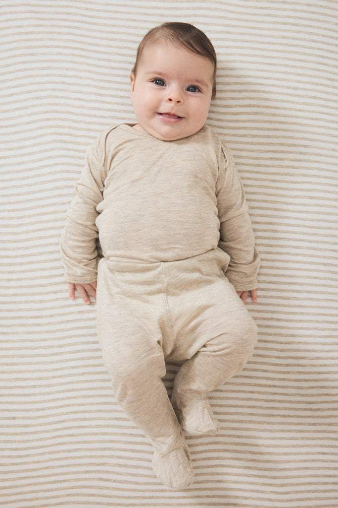 A baby lying on their back wearing a beige long-sleeve footed sleeper, photographed from a top-down view on a striped beige and white blanket.