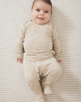 A baby lying on their back wearing a beige long-sleeve footed sleeper, photographed from a top-down view on a striped beige and white blanket.