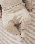 A close-up of a baby wearing a heathered beige footed sleeper, lying on a striped beige and white blanket.
