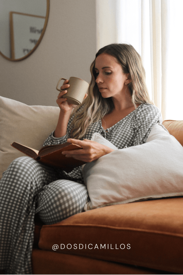 Woman in checkered pajamas reading a book and holding a mug on a couch. Features the instagram handle @dosdicamillos