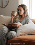 Woman in checkered pajamas reading a book and holding a mug on a couch. Features the instagram handle @dosdicamillos