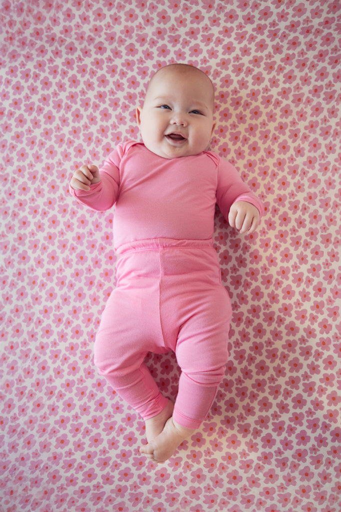 Smiling baby wearing solid pink long-sleeve top and pants lying on pink floral patterned blanket.