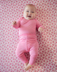 Smiling baby wearing solid pink long-sleeve top and pants lying on pink floral patterned blanket.
