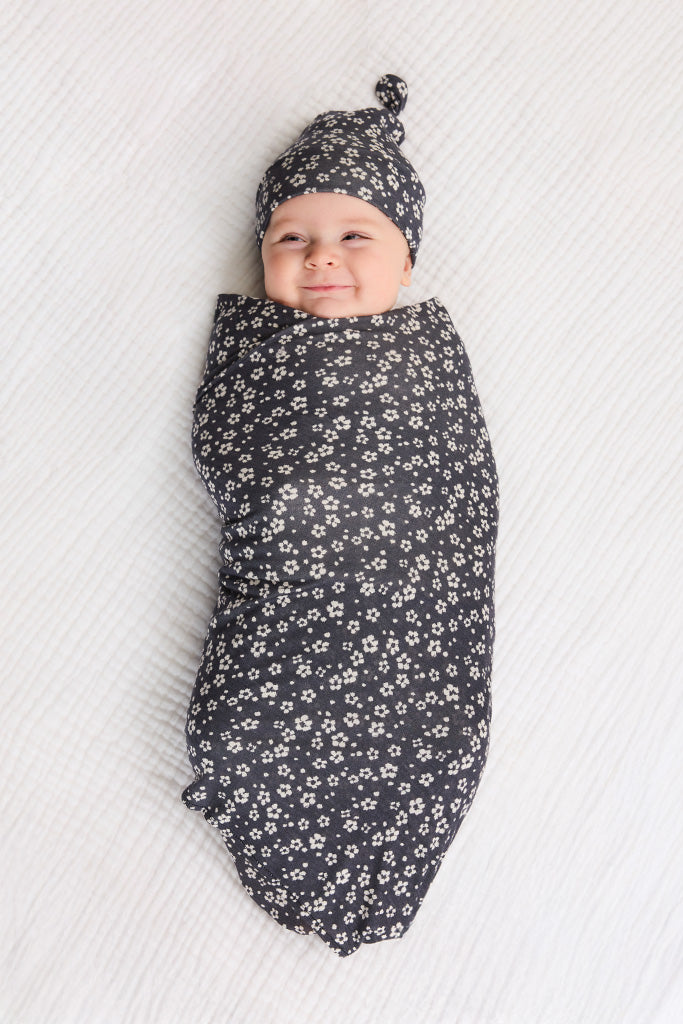 Baby swaddled in black fabric with white floral print and matching knotted hat on white textured surface.