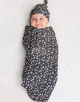 Baby swaddled in black fabric with white floral print and matching knotted hat on white textured surface.