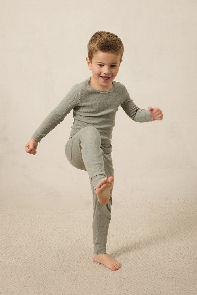 A boy wearing a mist pointelle sleep set, standing with one foot up on a neutral background