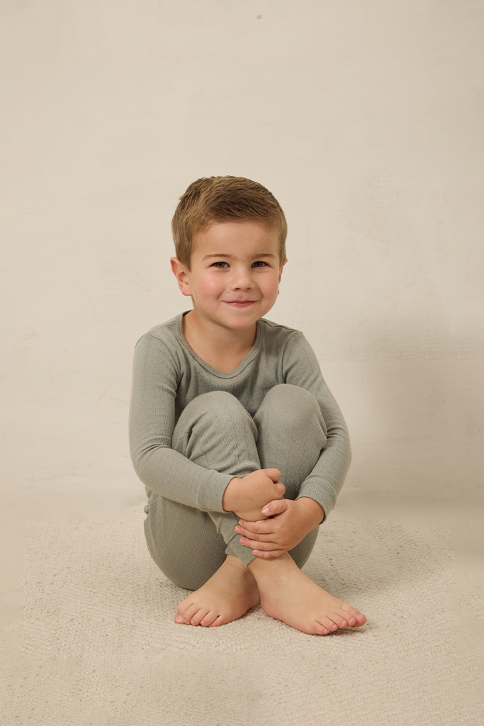 Young boy wearing a mist pointelle sleep set sitting on a beige surface with a plain background
