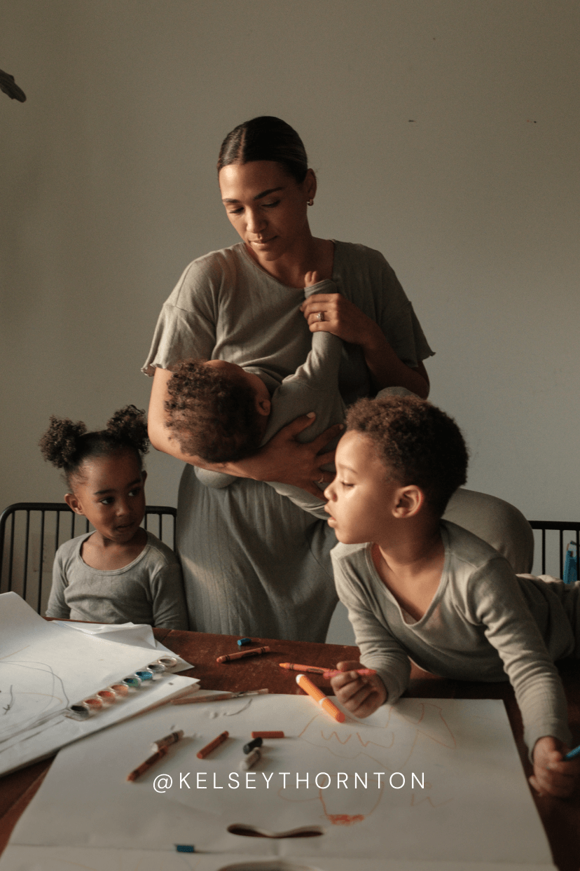 A woman holding a baby standing near two children sitting at a table with art supplies