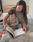 A woman and a child reading a book sitting on a bed in matching mist pointelle pajamas