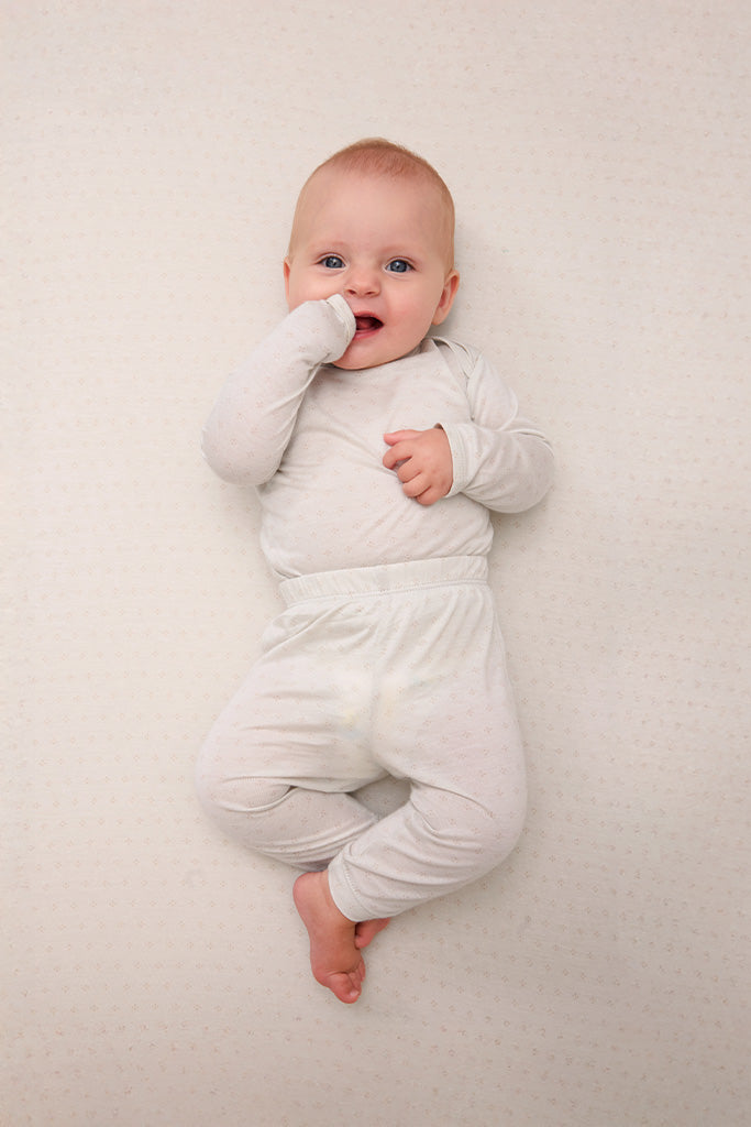 Baby wearing light beige long sleeve onesie and pants with subtle dot pattern lying on matching light beige textured surface.