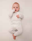 Baby wearing light beige long sleeve onesie and pants with subtle dot pattern lying on matching light beige textured surface.