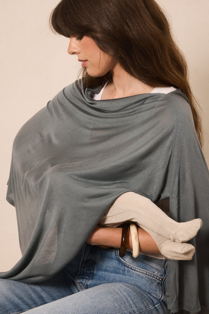 A woman wearing a draped sage green shawl or wrap over her shoulders and around her arms, with blue jeans and a white top visible. She is posed against a plain beige studio background.