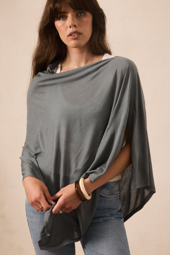 A woman wearing a draped, oversized gray top with wide sleeves and a loose silhouette, paired with blue jeans, standing against a plain beige background.
