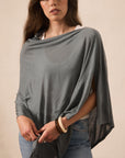 A woman wearing a draped, oversized gray top with wide sleeves and a loose silhouette, paired with blue jeans, standing against a plain beige background.