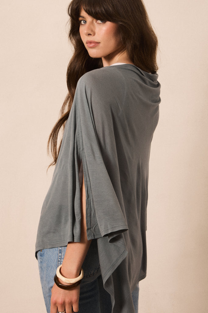 A woman wearing a draped, oversized gray top with wide sleeves and side slits, paired with blue jeans, photographed against a plain beige studio background.