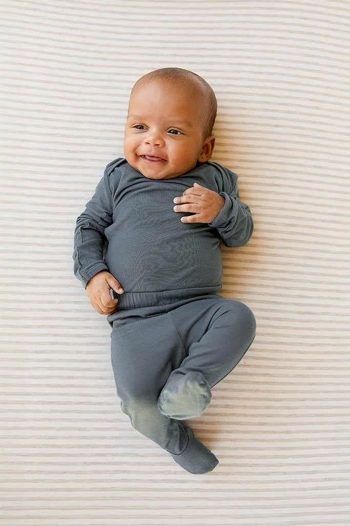 Baby wearing long-sleeve dark gray footed pajama lying on a beige and white striped surface.
