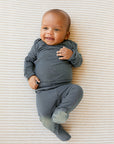 Baby wearing long-sleeve dark gray footed pajama lying on a beige and white striped surface.