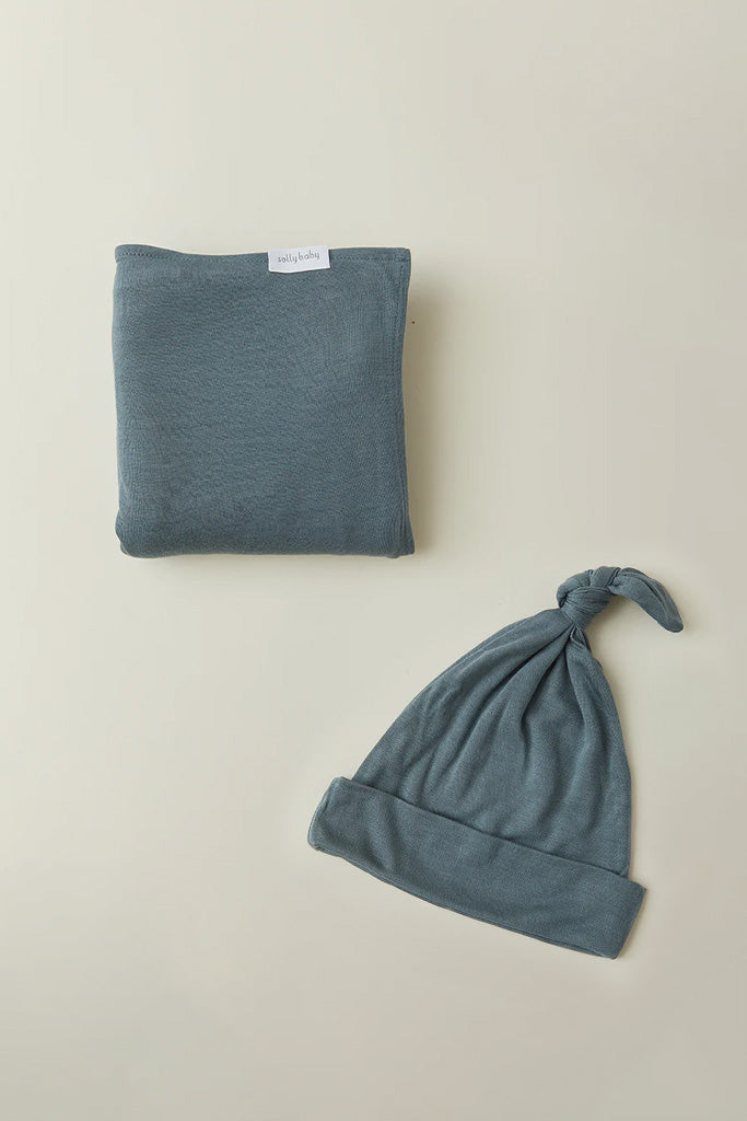 Blue-gray baby hat with top knot and matching folded blanket with a small white brand label.