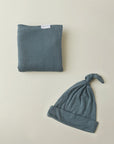 Blue-gray baby hat with top knot and matching folded blanket with a small white brand label.