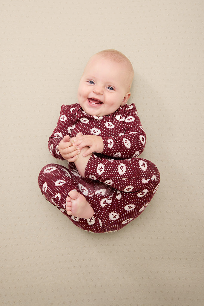 Smiling baby in maroon long-sleeve onesie with white apple and dot pattern sitting on beige surface.