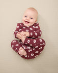Smiling baby in maroon long-sleeve onesie with white apple and dot pattern sitting on beige surface.