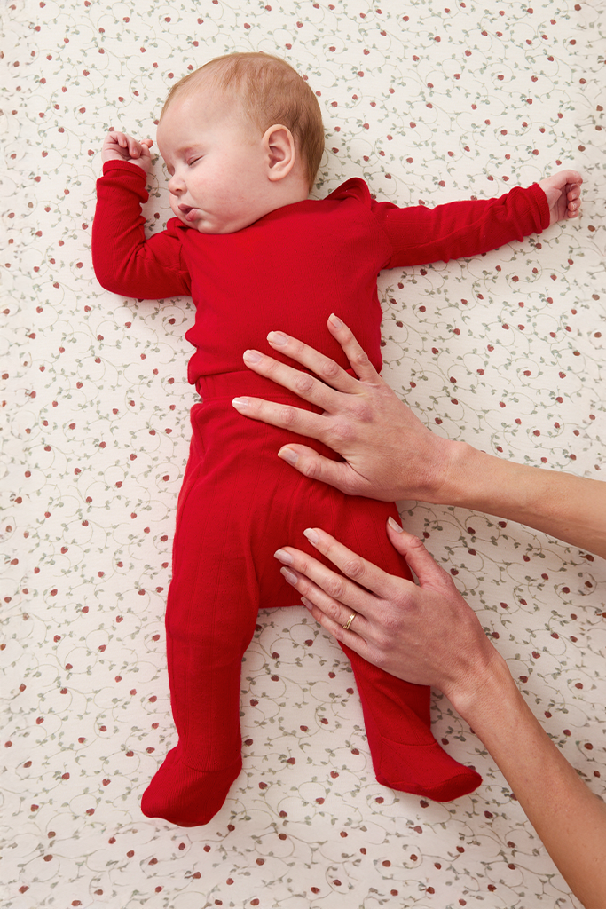 Baby in a red sleep set lying on a patterned surface with adult hands gently touching the baby.
