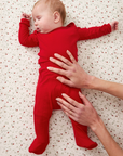 Baby in a red sleep set lying on a patterned surface with adult hands gently touching the baby.