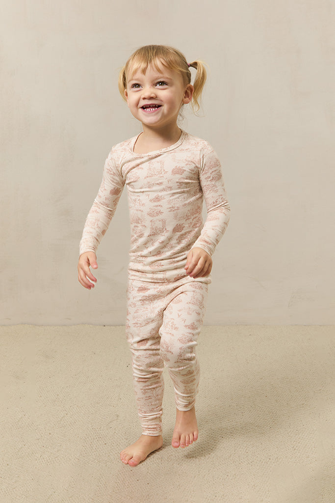 Toddler girl wearing beige long-sleeve top and pants set with light brown scenic pattern, standing barefoot on beige carpet.