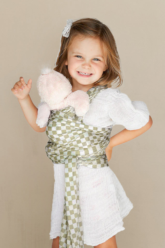 Smiling girl in white dress wearing green and white checkered baby wrap holding light pink plush toy.