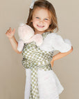Smiling girl in white dress wearing green and white checkered baby wrap holding light pink plush toy.