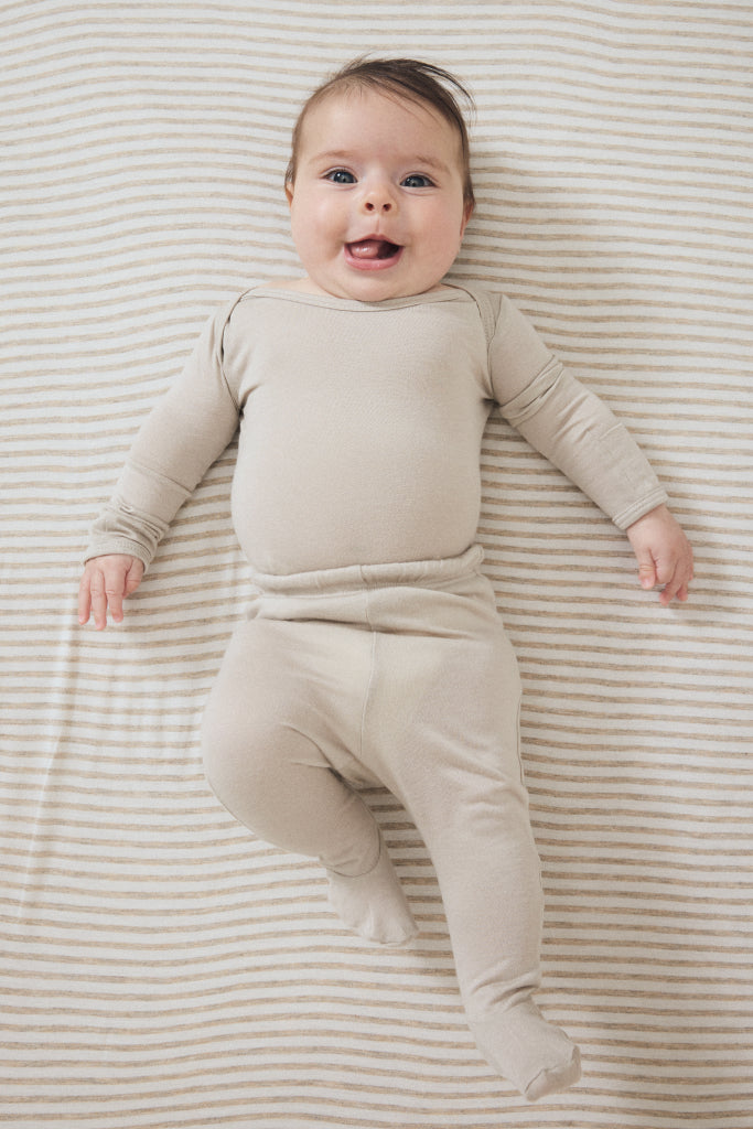 A smiling baby lying on their back on a beige and white striped blanket, wearing a matching beige long-sleeve footed sleeper.