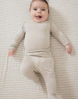 A smiling baby lying on their back on a beige and white striped blanket, wearing a matching beige long-sleeve footed sleeper.