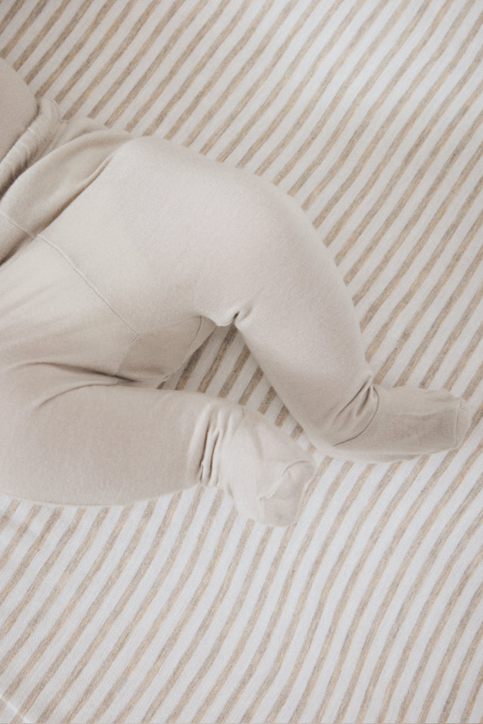 A close-up of a baby's legs and feet wearing a soft, light beige footed sleeper or bootie, lying on a striped beige and white mattress.