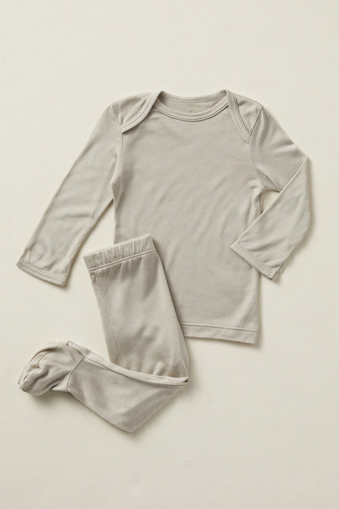 A two-piece baby pajama set in a light oatmeal gray, featuring a long-sleeve top with shoulder snaps and matching footed pants, laid flat on a neutral cream background.