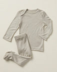 A two-piece baby pajama set in a light oatmeal gray, featuring a long-sleeve top with shoulder snaps and matching footed pants, laid flat on a neutral cream background.