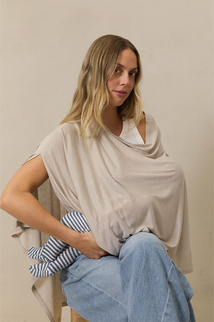 Beige nursing cover worn by woman holding a baby dressed in blue and white striped outfit while seated on a wooden stool.