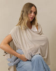 Beige nursing cover worn by woman holding a baby dressed in blue and white striped outfit while seated on a wooden stool.