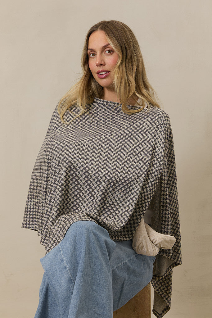 Black and beige houndstooth poncho on model sitting wearing light blue jeans against beige background
