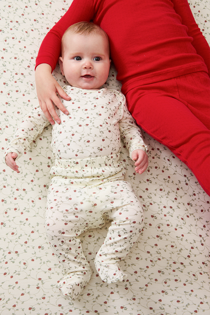 Baby in a strawberry outfit lying on a patterned blanket with an arm from big sib visible