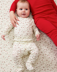 Baby in a strawberry outfit lying on a patterned blanket with an arm from big sib visible