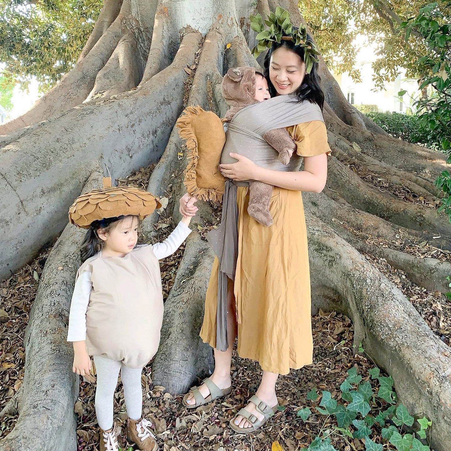 Woman in mustard dress with leafy headband carrying baby in gray wrap, and child in beige acorn costume holding a pillow.